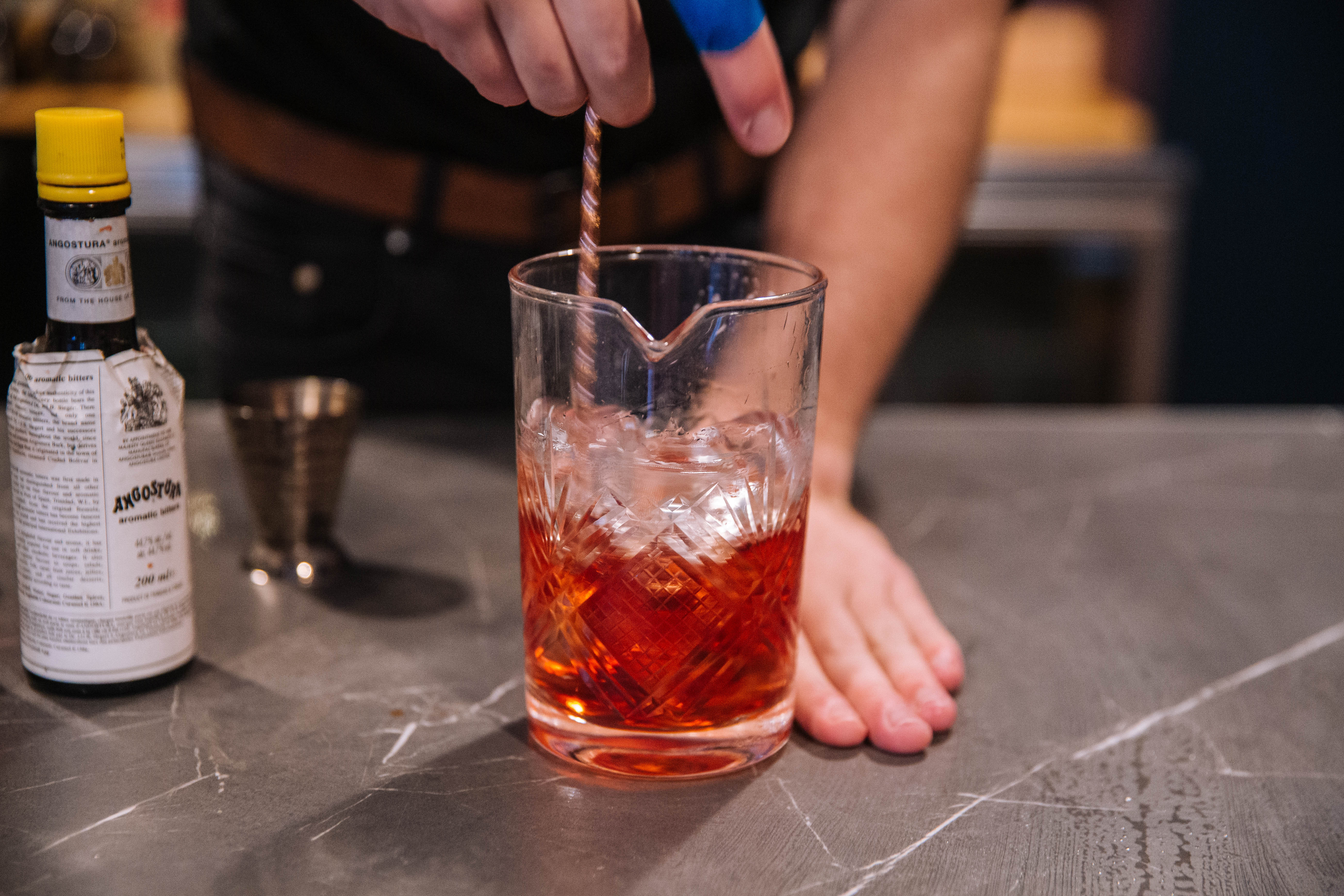 Hand stirring an orange-colored cocktail with Angostura bitters in the background